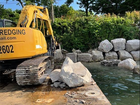 A yellow excavator places large light-colored rocks along a shoreline to build a retaining wall.