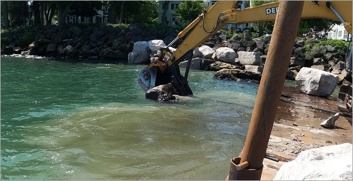 A yellow excavator places large boulders into the water along a rocky shoreline, causing a plume of sediment.