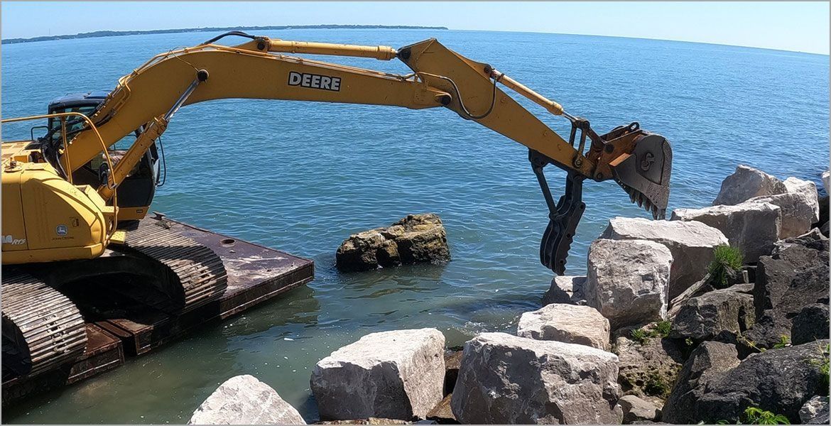A yellow excavator on a floating platform places large boulders along a shoreline into the water.