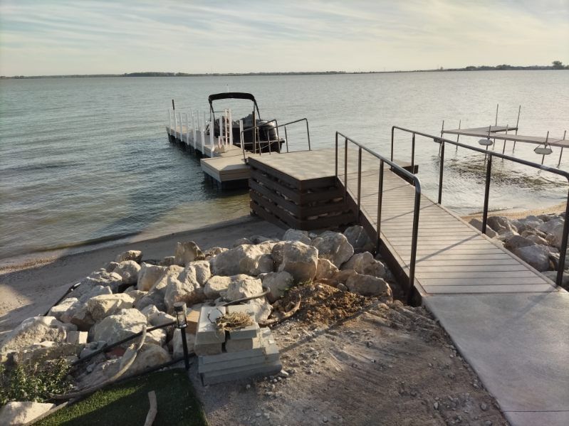 A boat docked at a pier extending into a calm lake, with a wooden walkway and rock-lined shore in the foreground.