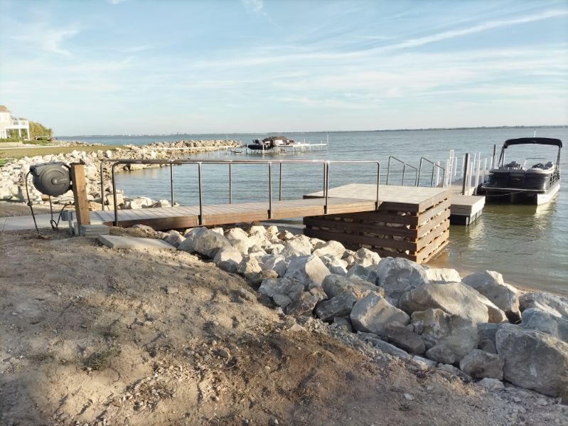A wooden dock with metal railings extends into a lake from a rocky shore, with a pontoon boat moored nearby.