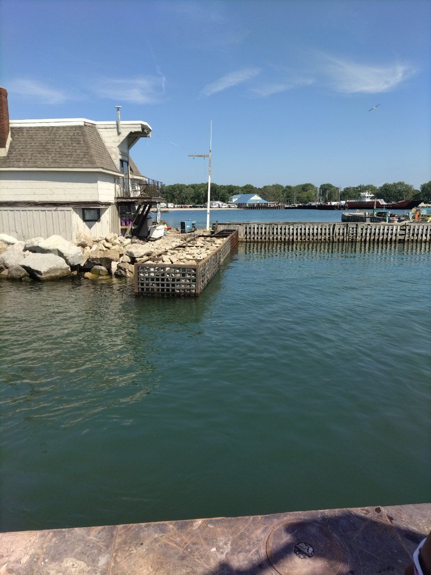 A waterfront view featuring a light-colored building with a rocky pier extending into calm, blue water under a clear sky.