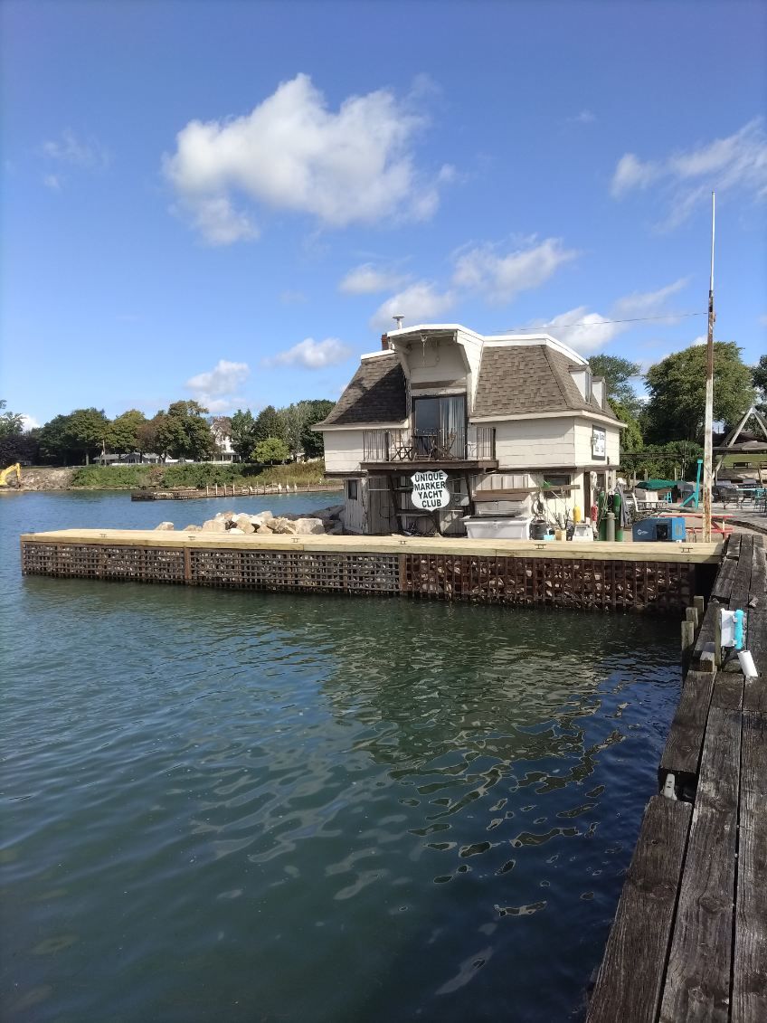 A small, rustic house sits on a stone pier extending into a calm body of water under a bright blue sky.