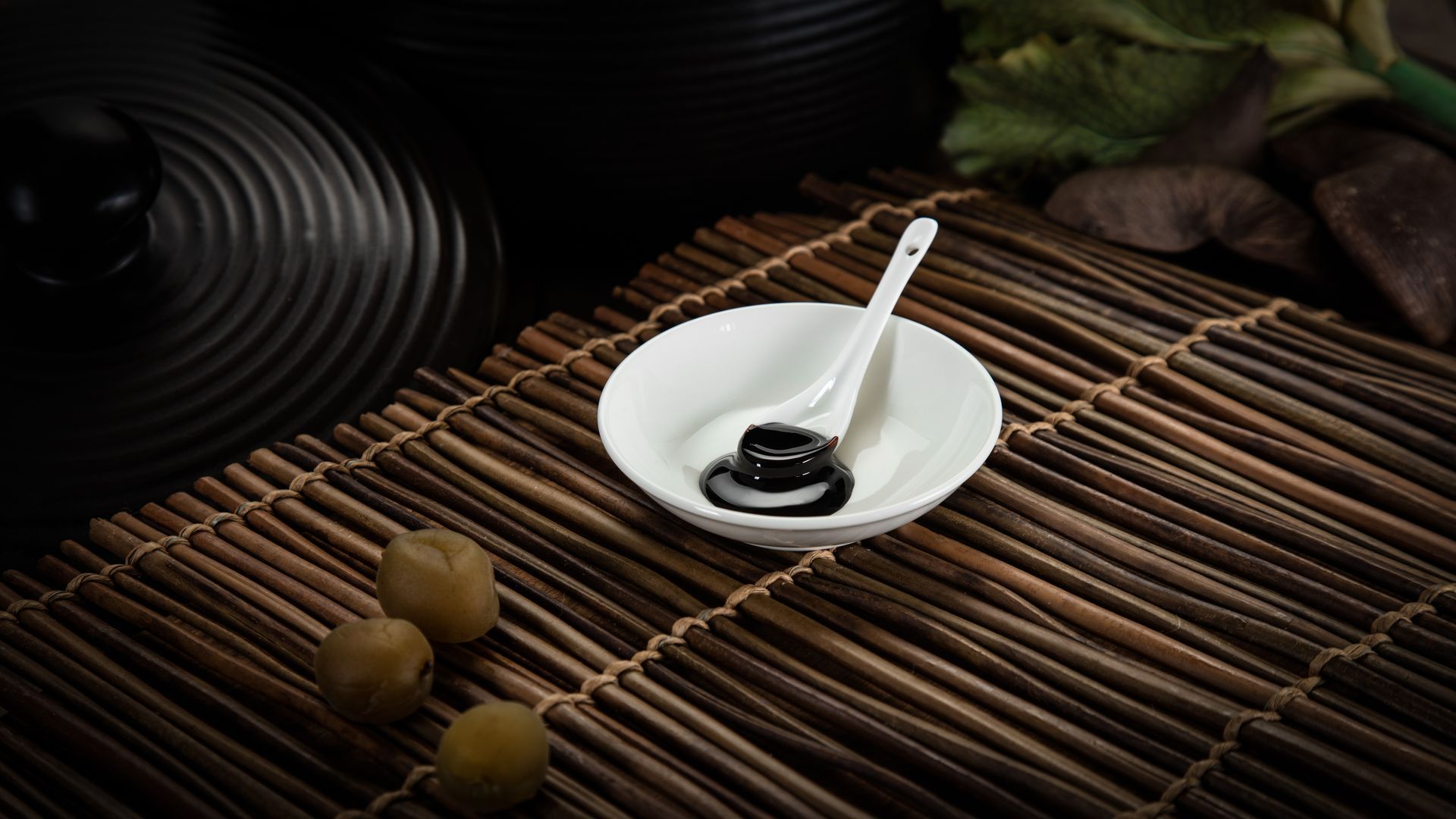白色碗盛著黑色醬汁,湯匙放在竹蓆上,背景中有橄欖和深色盤子。