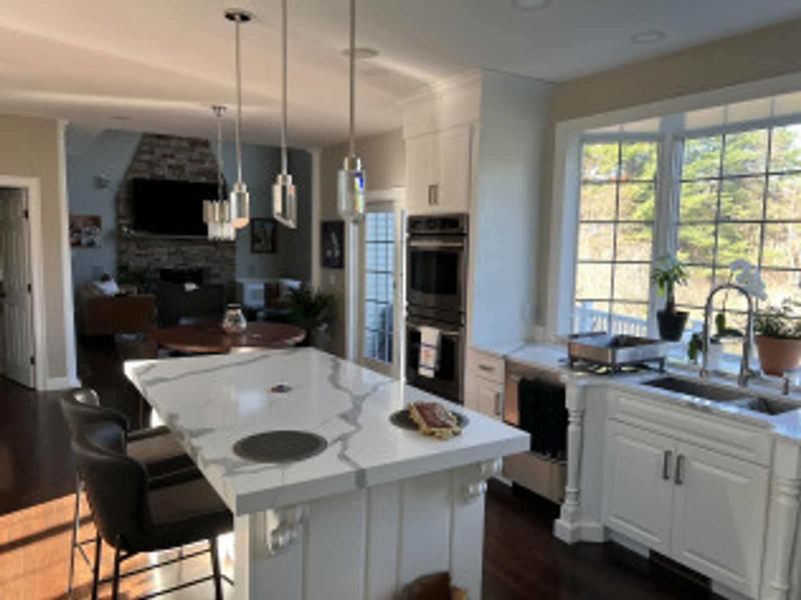 Spacious kitchen with white cabinets, island, and sink by a large window overlooking trees. Living room in background.