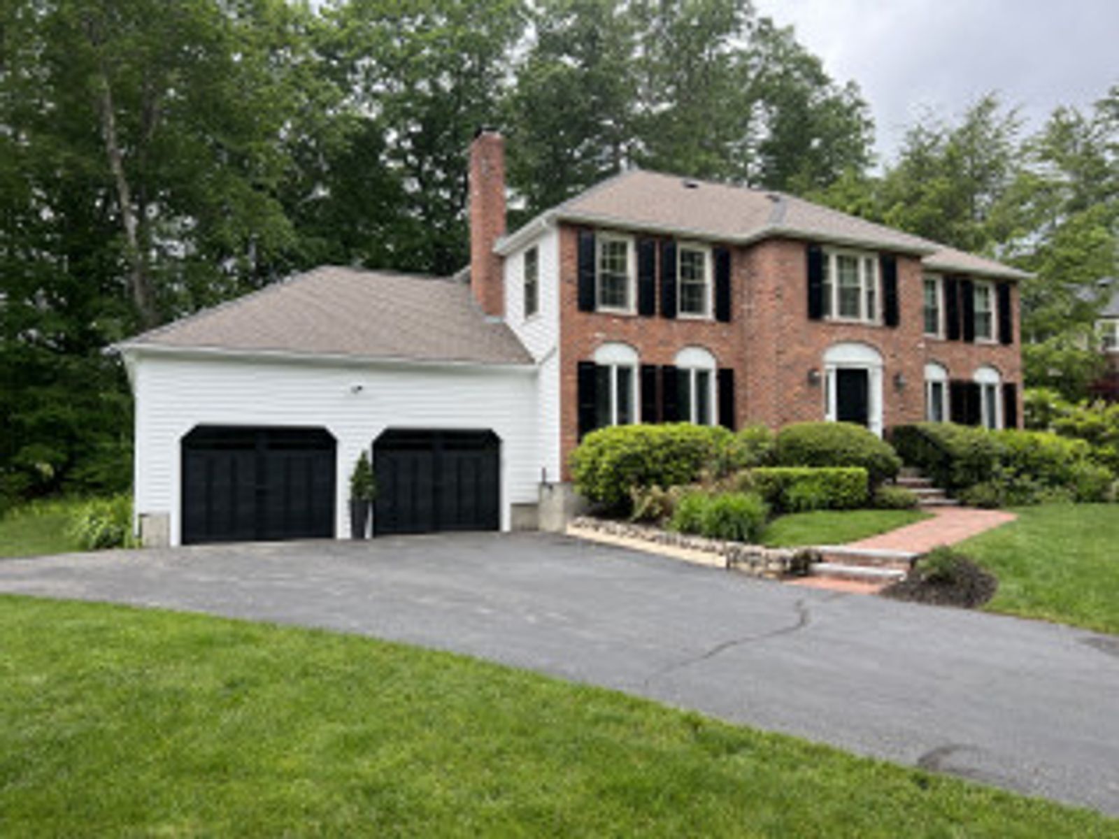 Brick house with black shutters, two-car garage, and asphalt driveway. Lush green lawn and trees.