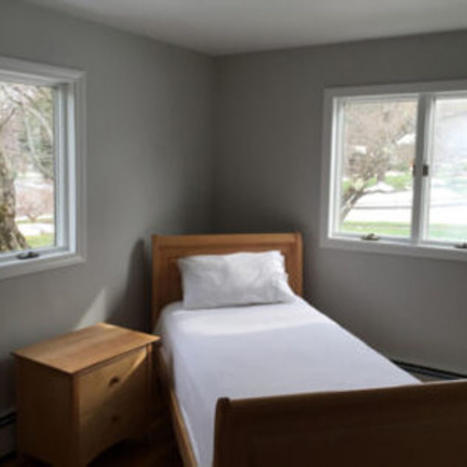 Bedroom with single bed, bedside table, and two windows. Gray walls and natural light.