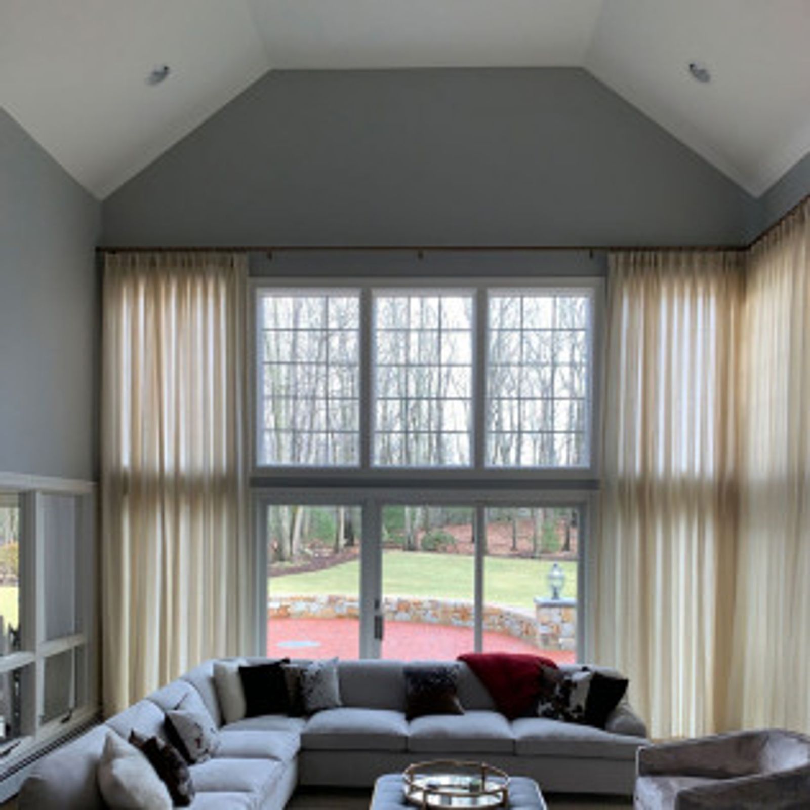 Living room with high ceilings, large windows, and cream-colored curtains. A gray sectional sofa is visible.