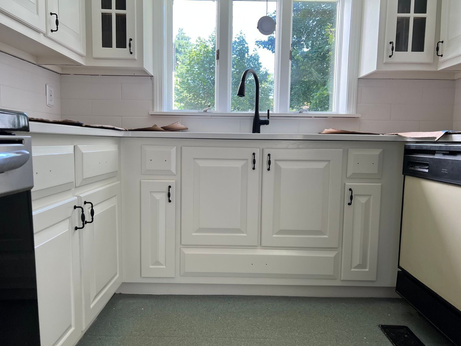 White kitchen cabinets with black hardware and faucet; sink in front of window.