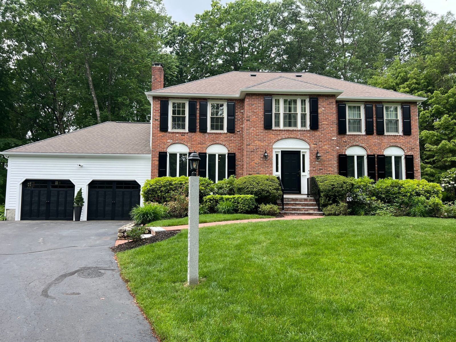 Two-story brick house with black shutters, attached garage, and green lawn. Asphalt driveway.