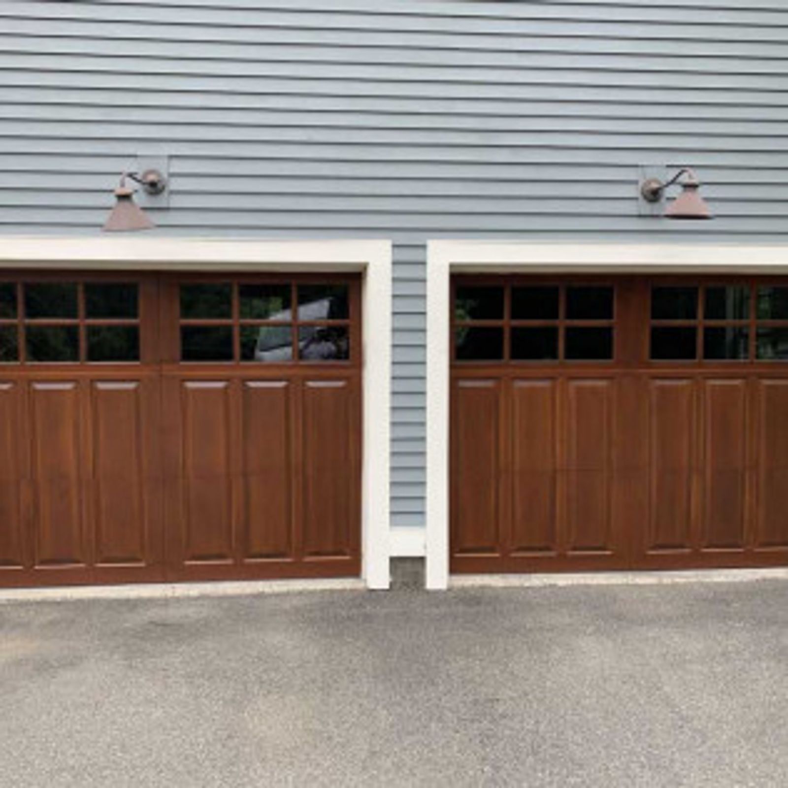 Two brown wooden garage doors with windows, flanked by gray siding, white trim, and copper lights.