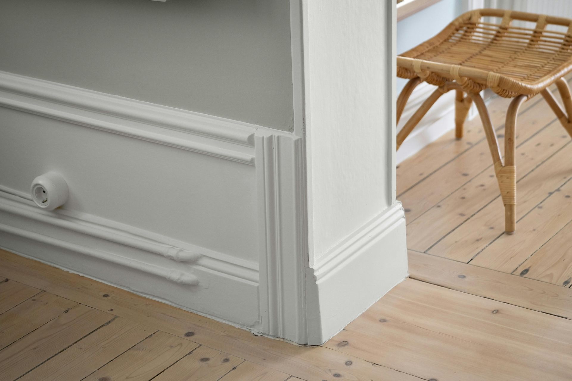 White wainscoting and molding meet light wood flooring. A woven stool sits in the background.