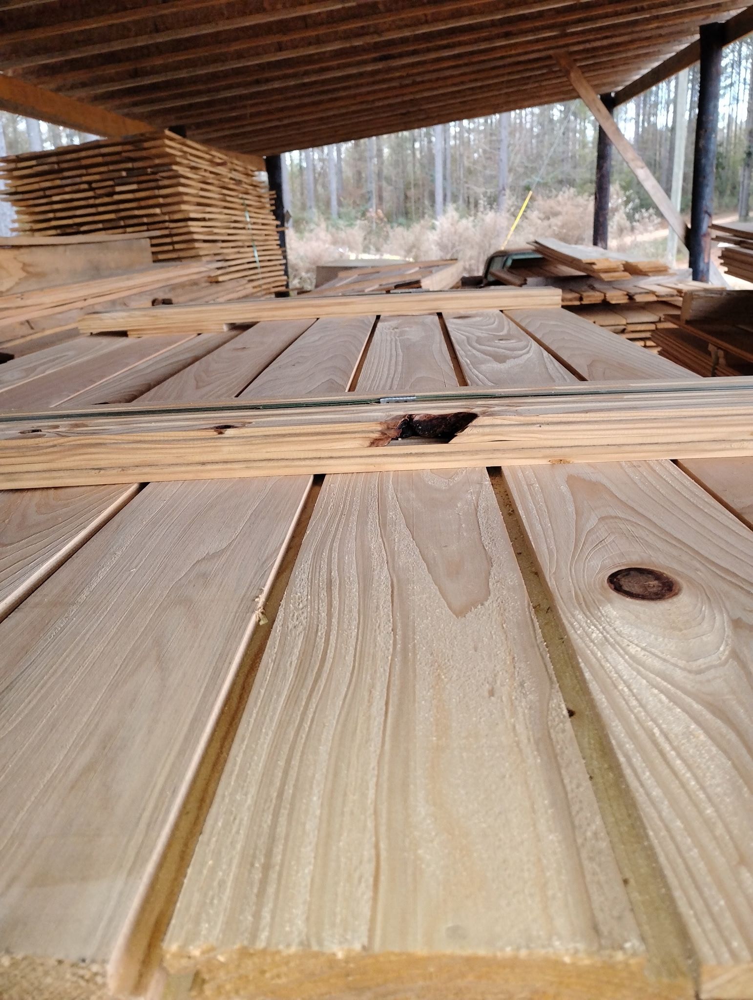 Wooden planks stacked in a lumberyard, partially covered by a roof.