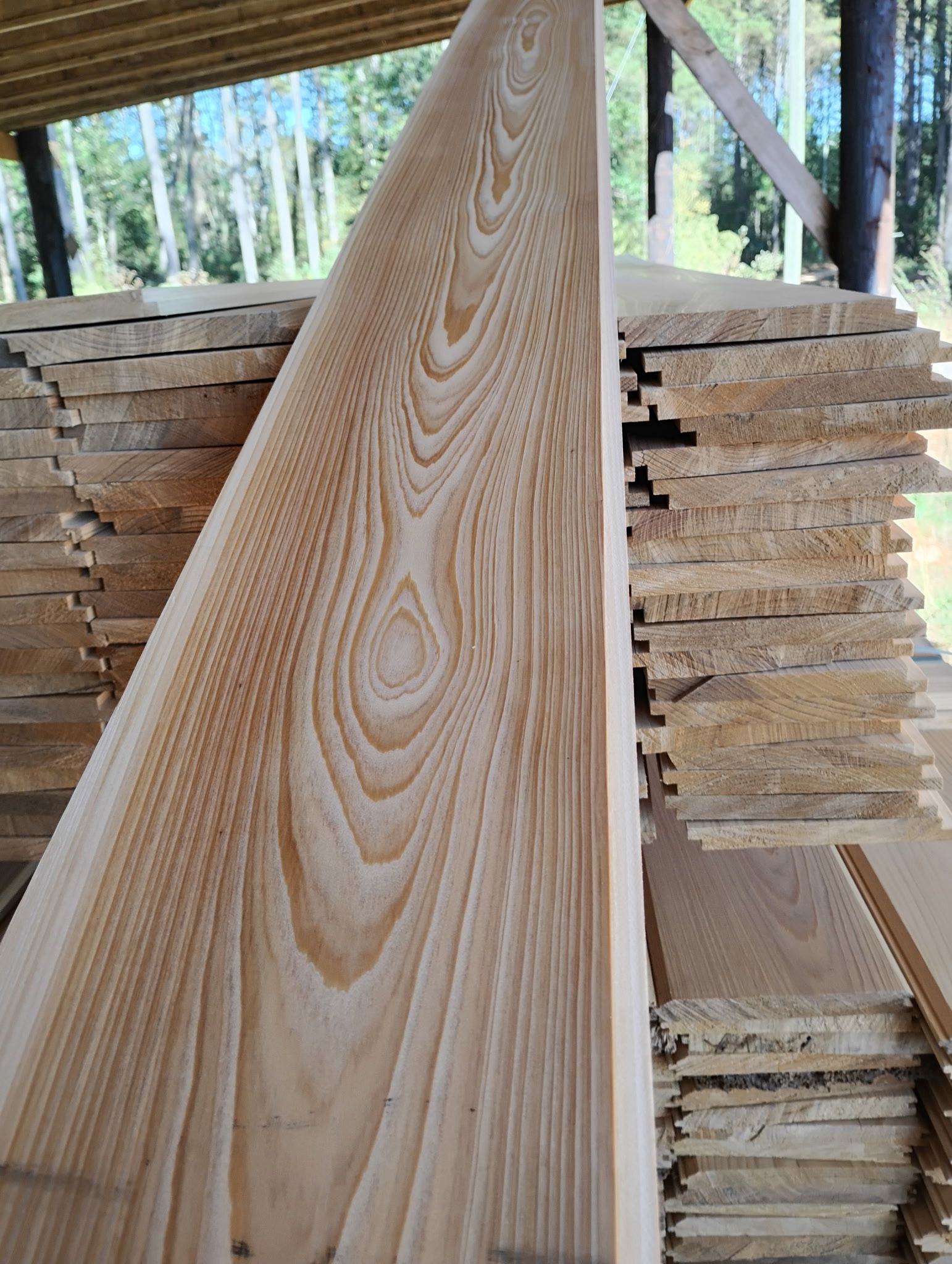 Wooden planks stacked outdoors, showing natural grain patterns, with a single plank in focus.