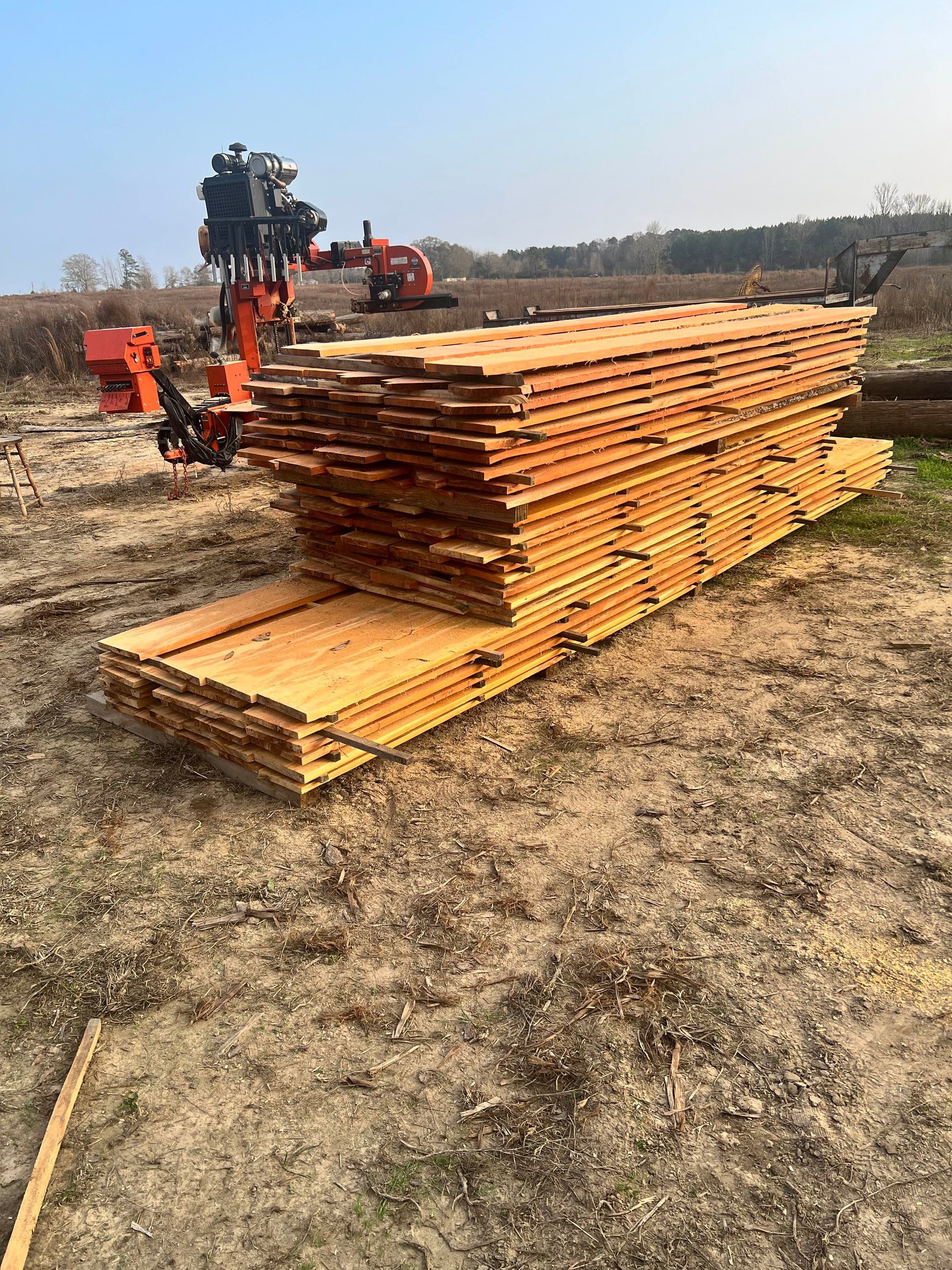 Stack of fresh-cut lumber in a field with logging equipment in the background. Brown wood, outdoor setting.