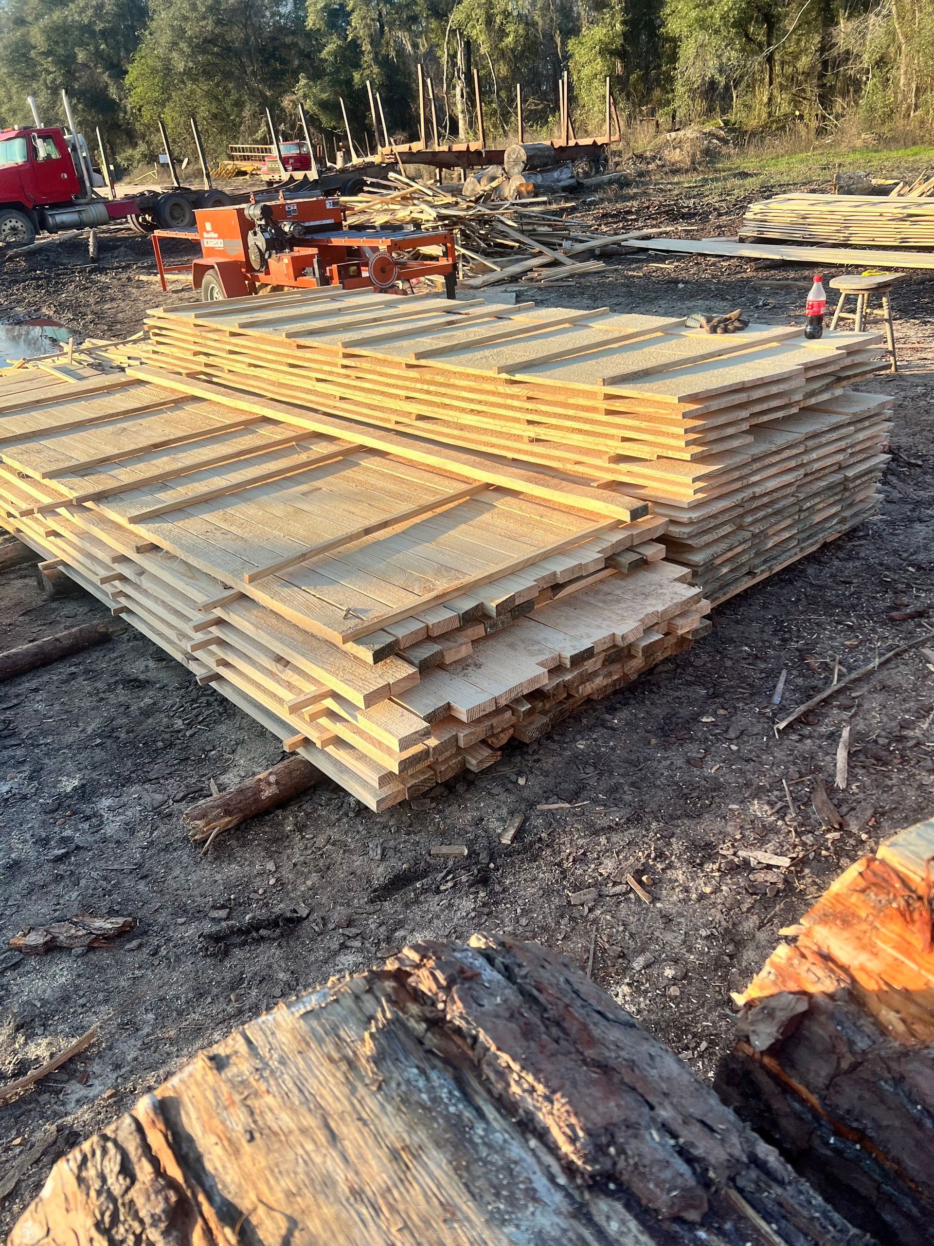 Stack of freshly-cut wood planks at a lumber mill, with machinery and red truck in the background.