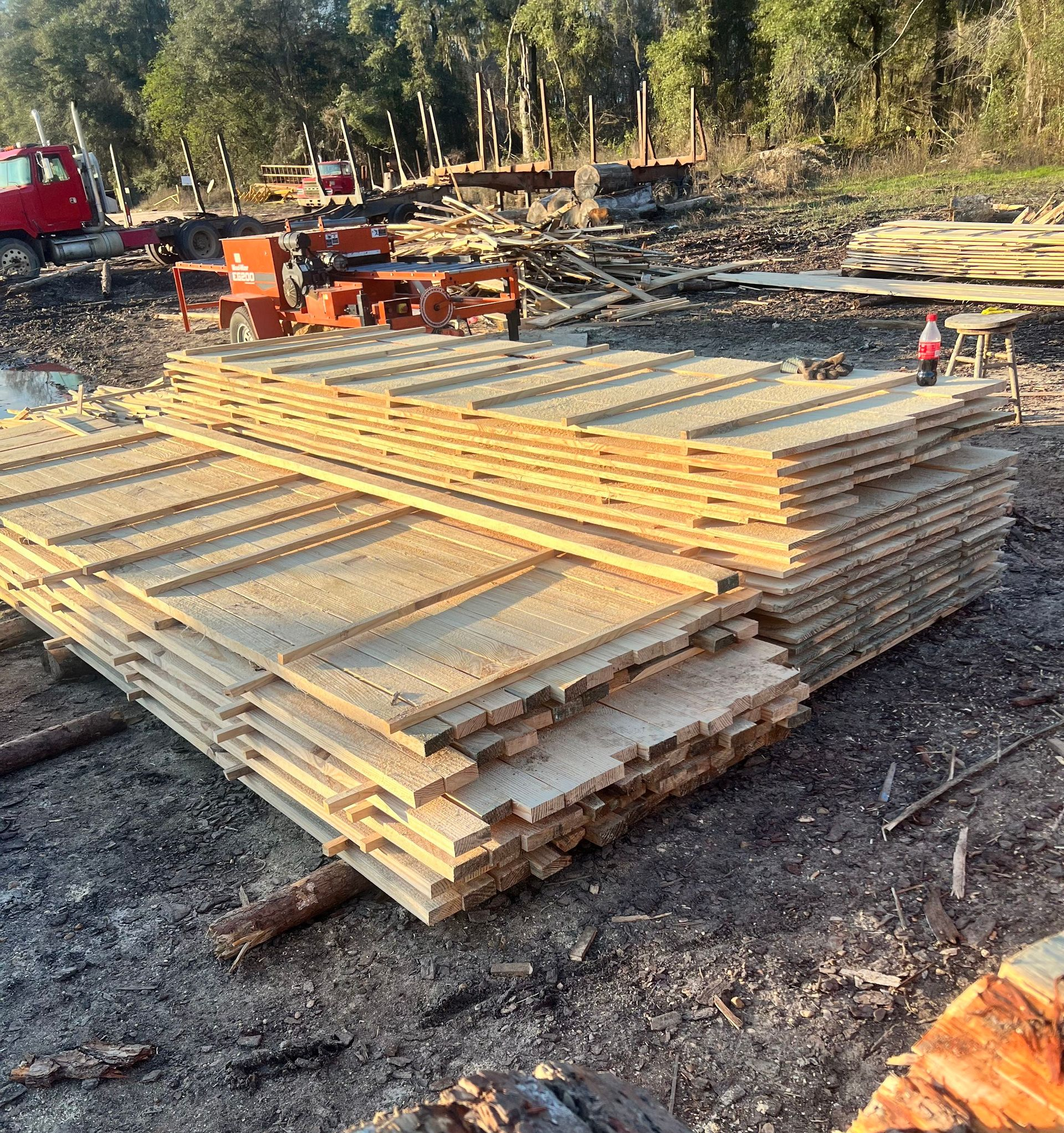 Stack of freshly cut wooden planks at a lumber mill, with machinery and logs in the background.