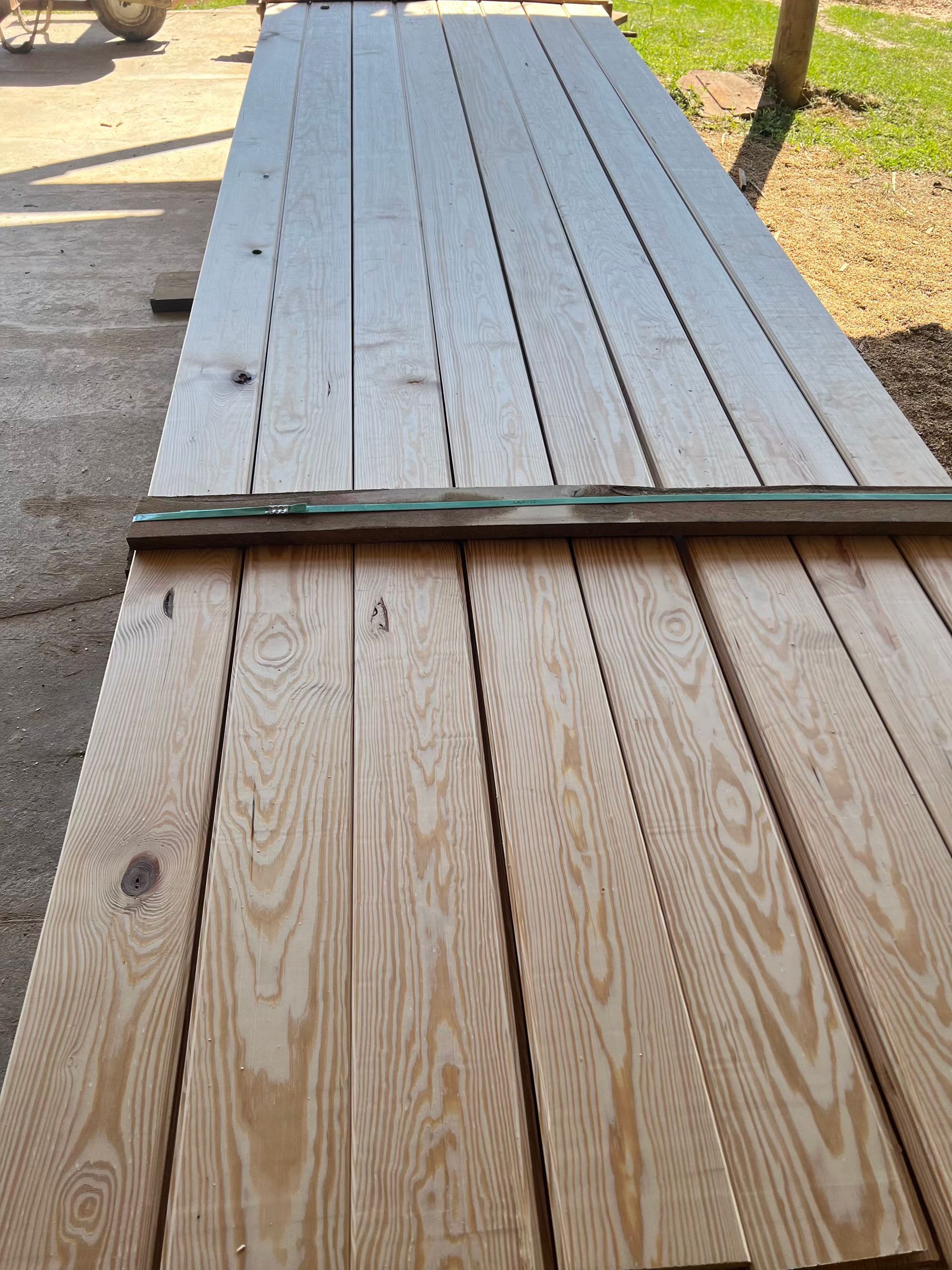 Wooden planks stacked outdoors, light brown with visible wood grain, secured by a green board.