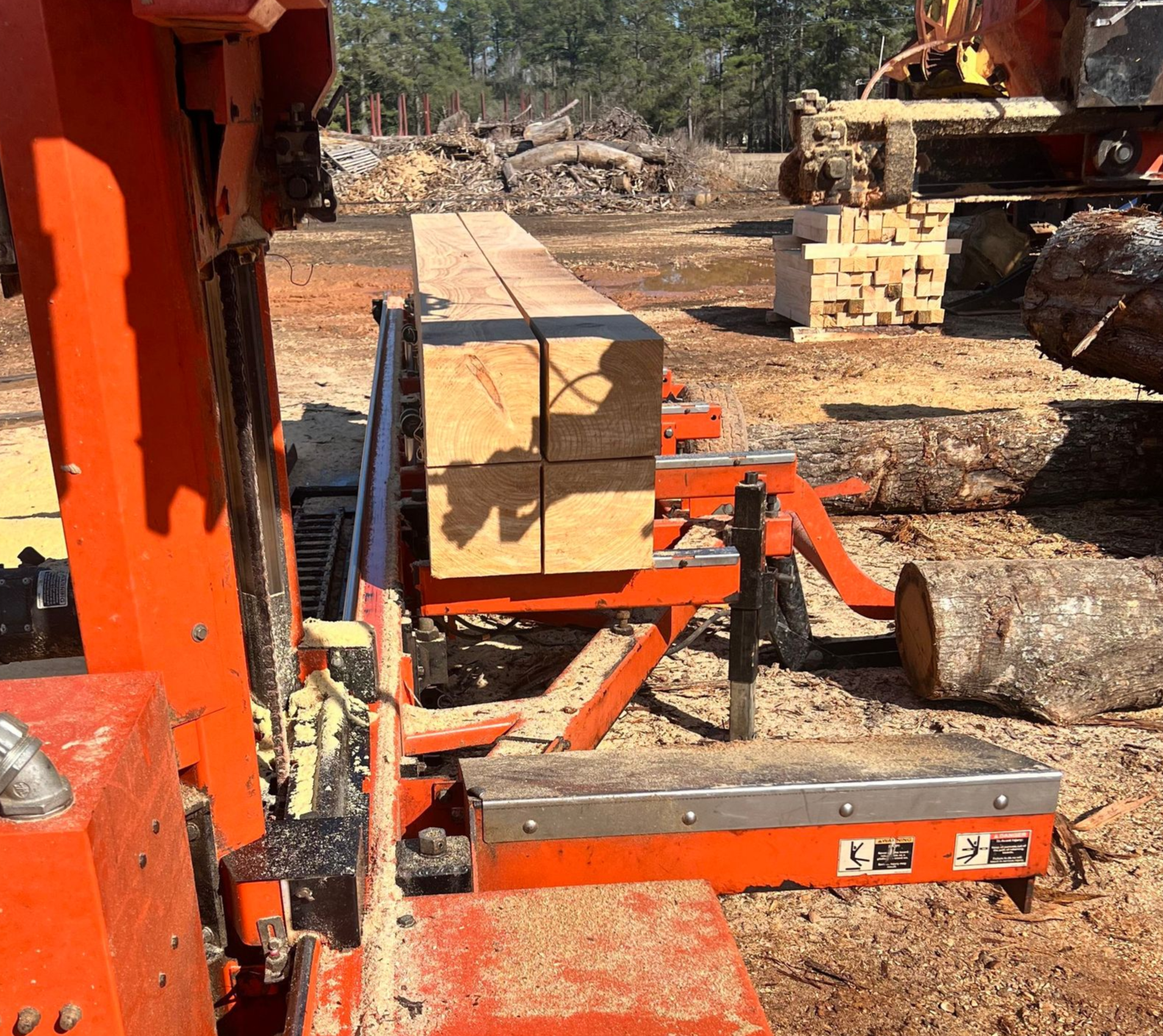 Lumber being cut on an orange sawmill outside. Saw is cutting through wood blocks. Wood shavings on the ground.