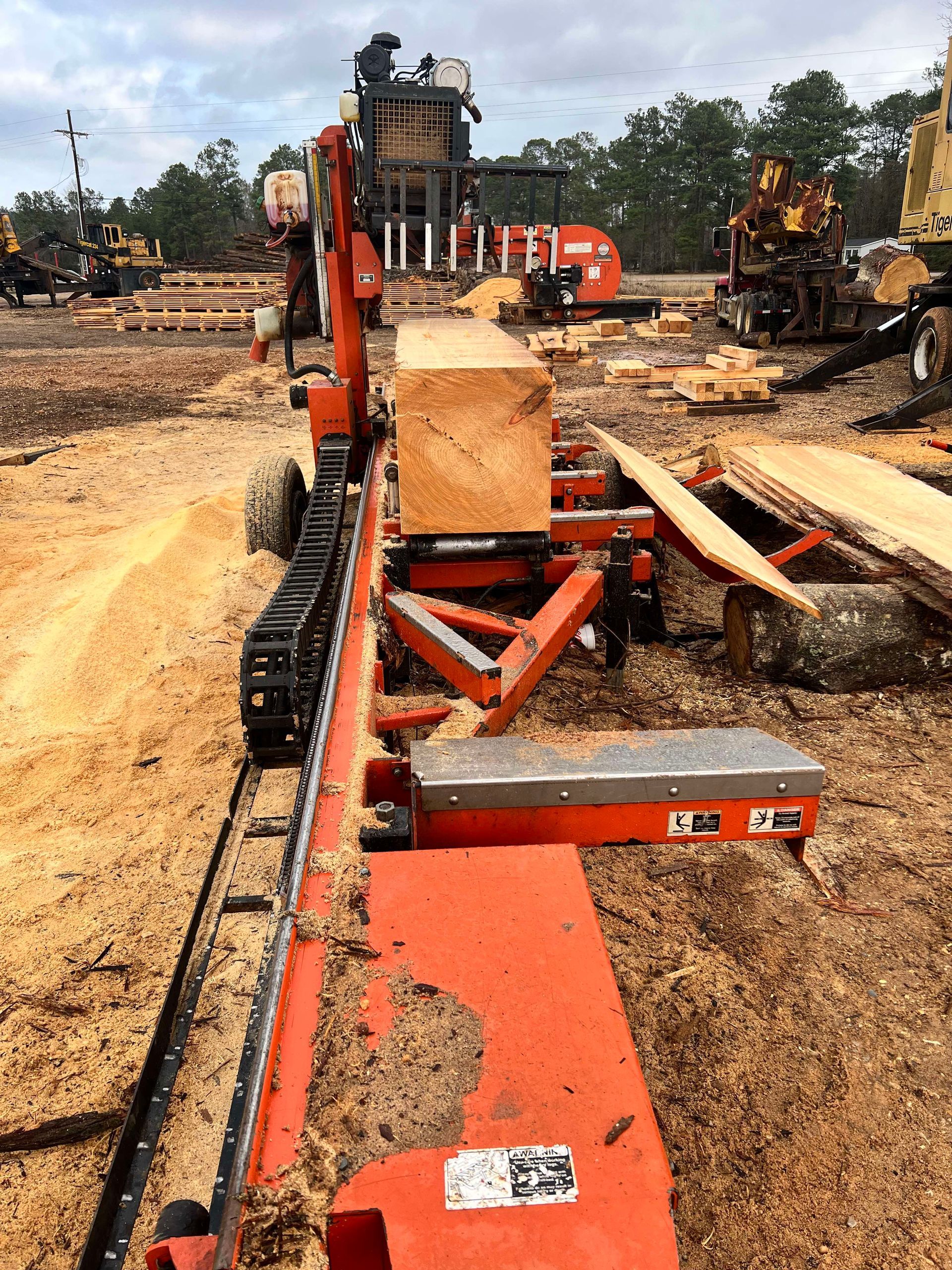 Orange sawmill cutting a large log into lumber outdoors; sawdust on the ground.