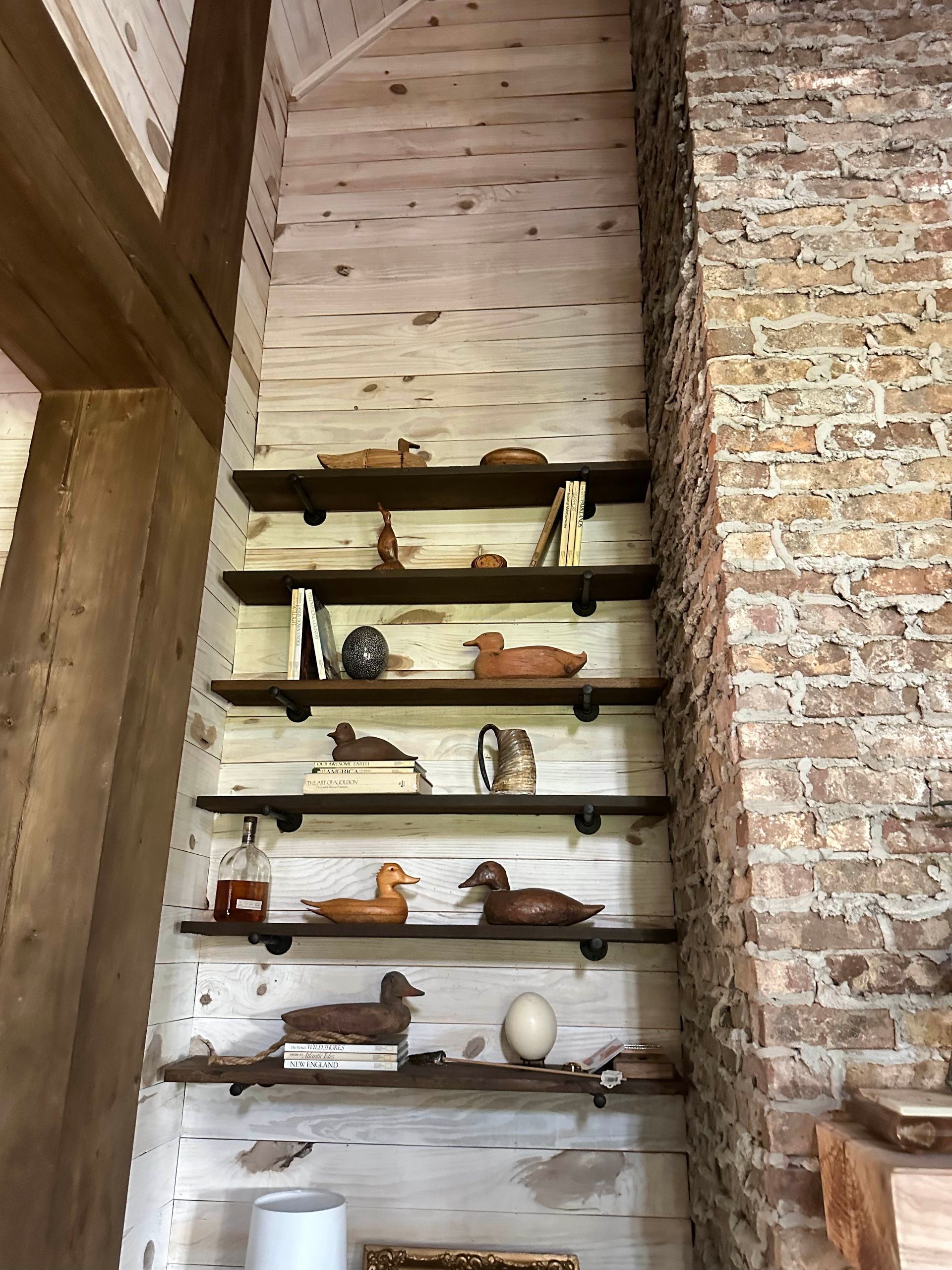 Wooden shelves against a white shiplap wall hold duck decoys and books. A brick wall is on the right.