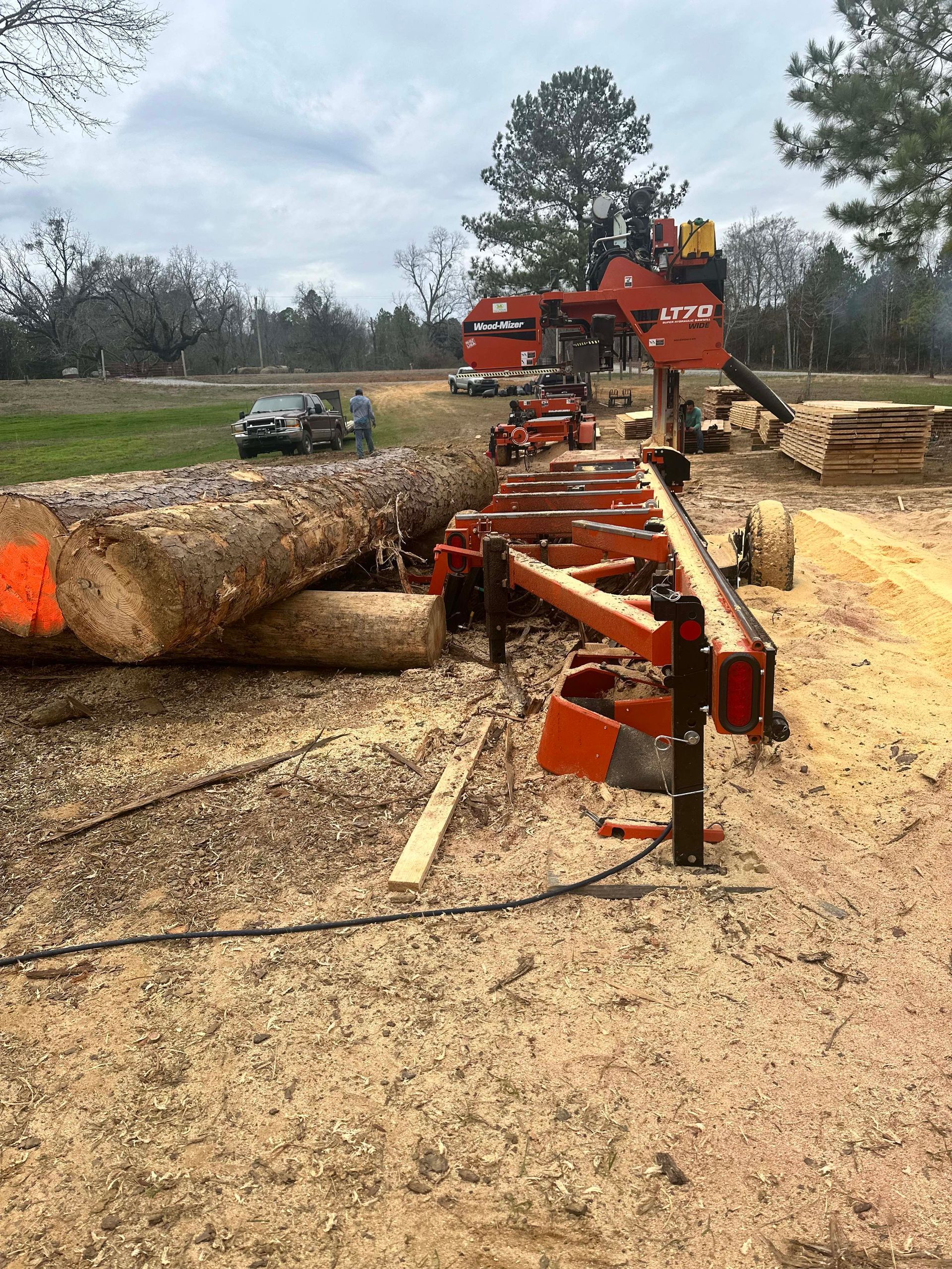 A sawmill cutting a large log into planks outdoors. Sawdust covers the ground.