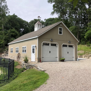 Beige detached garage with white trim, double doors, and gravel driveway beside a grassy yard and trees.