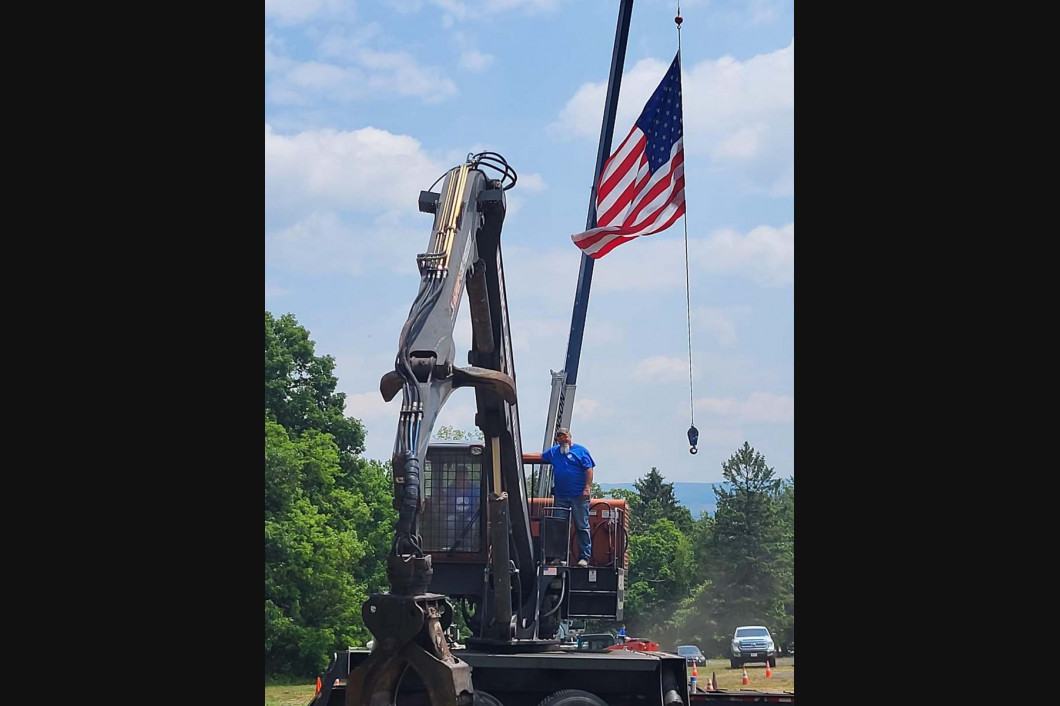 Worker on a truck-mounted lift beside a large American flag hanging from a tall pole.