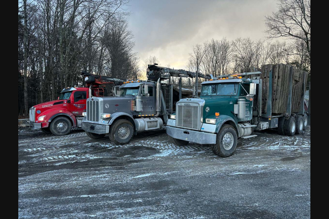 Three semi trucks parked on a snowy lot at dusk, including red, gray, and teal cabs.