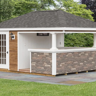 Small stone-and-white outdoor pavilion with a shingled roof beside a paved path and lawn