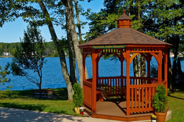 Wooden gazebo by a lakeshore, surrounded by trees and green grass on a sunny day