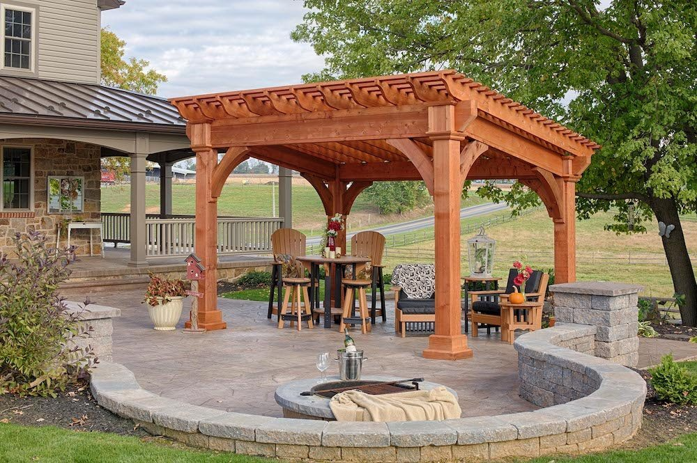 Wooden pergola gazebo with seating around a stone fire pit in a landscaped backyard