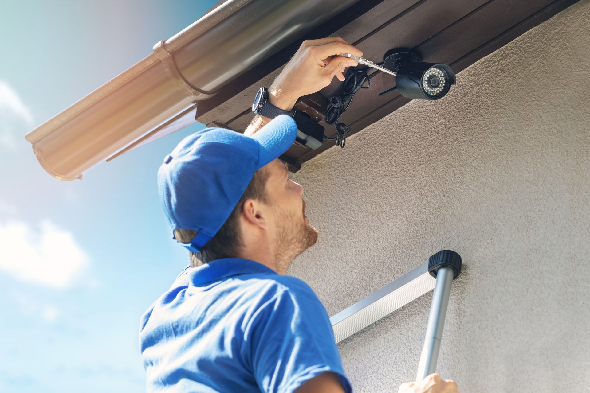 Man in blue uniform installing a security camera on a building exterior, holding a screwdriver.
