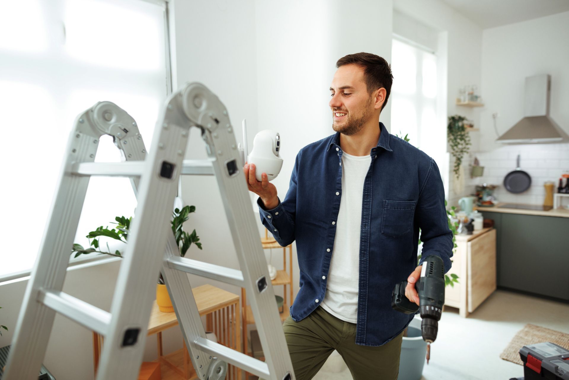 Man smiling, holding a security camera, near a ladder with a drill in hand; indoors, kitchen in background.