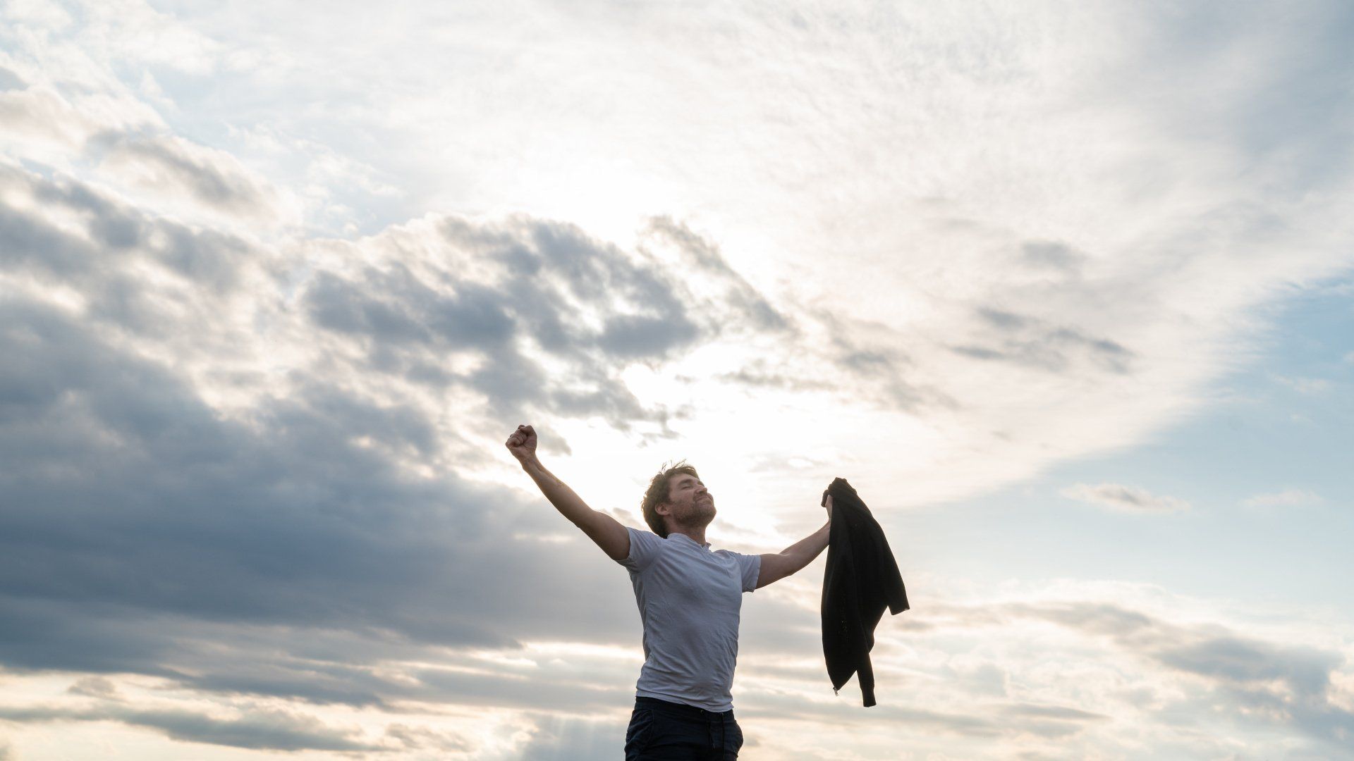 Een persoon staat met opgeheven armen tegen een uitgestrekte, bewolkte hemel en houdt een jas in één hand.