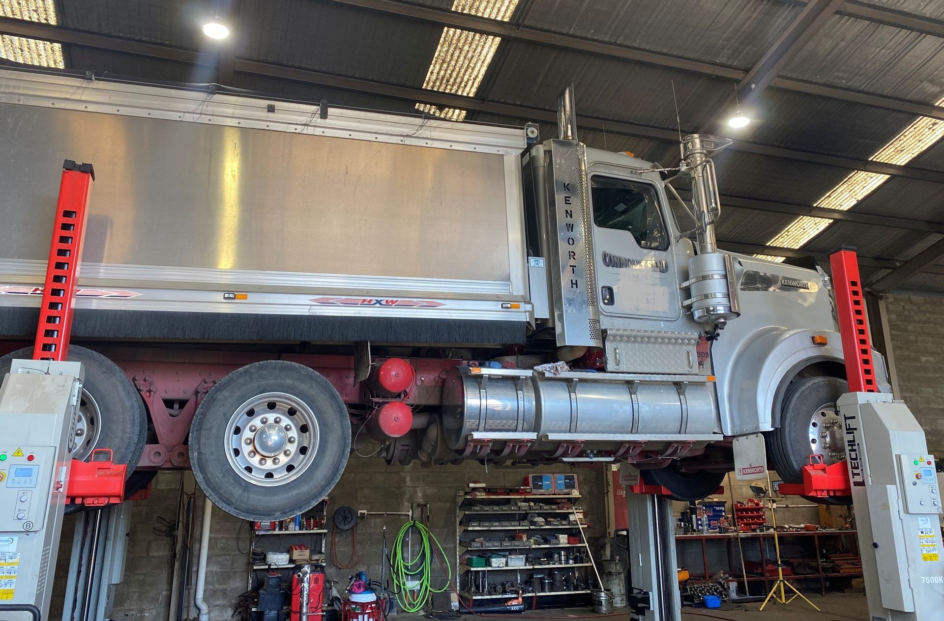 A silver truck raised on a lift in a repair shop; red lift arms, tools visible.