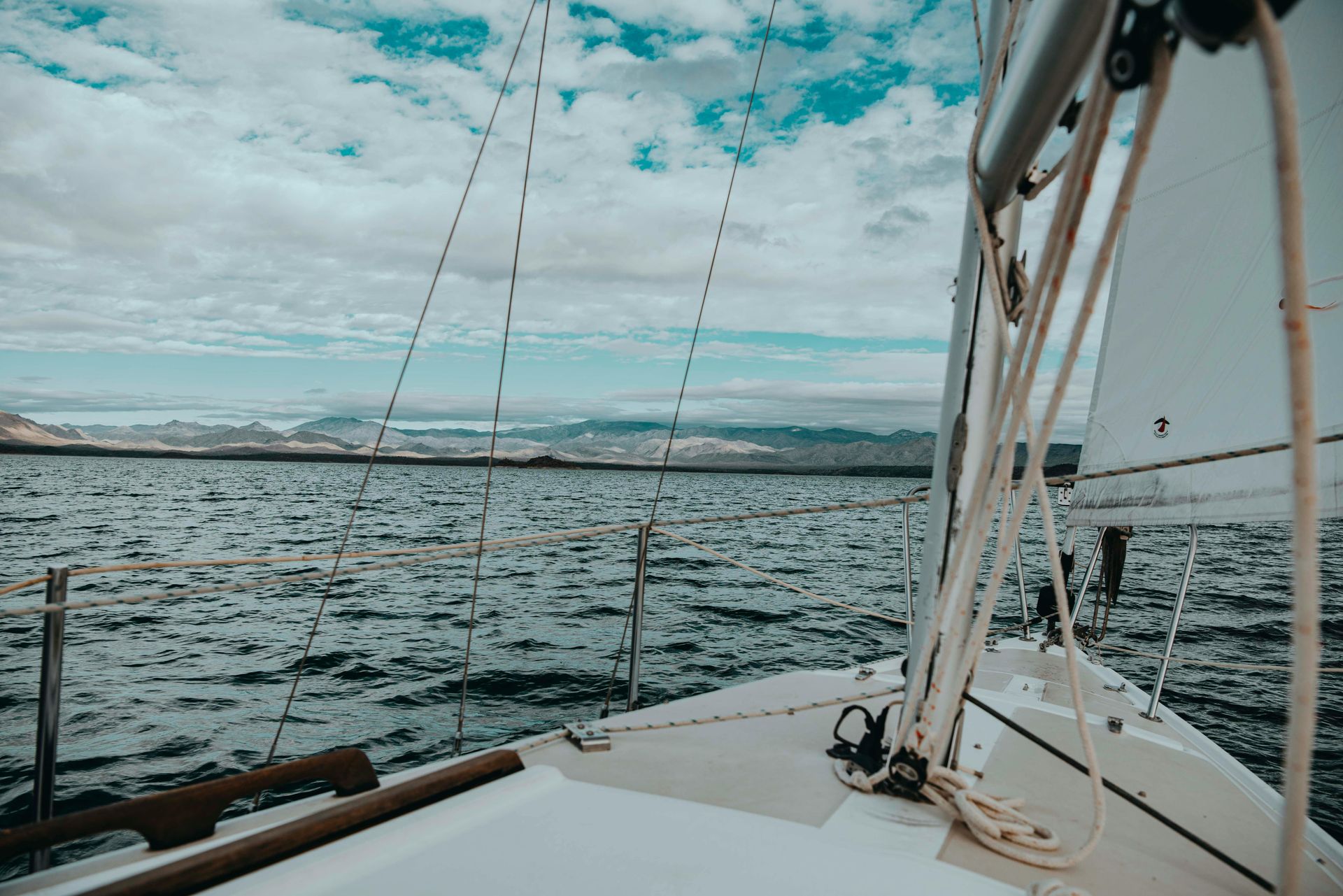 View from a sailboat deck over choppy water toward distant mountains under a cloudy sky