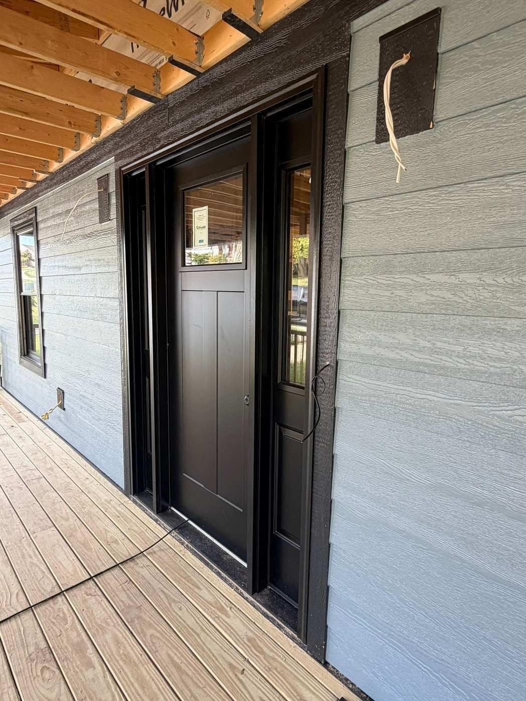 Newly installed black entry door with glass sidelites on a covered wood porch, viewed from the side