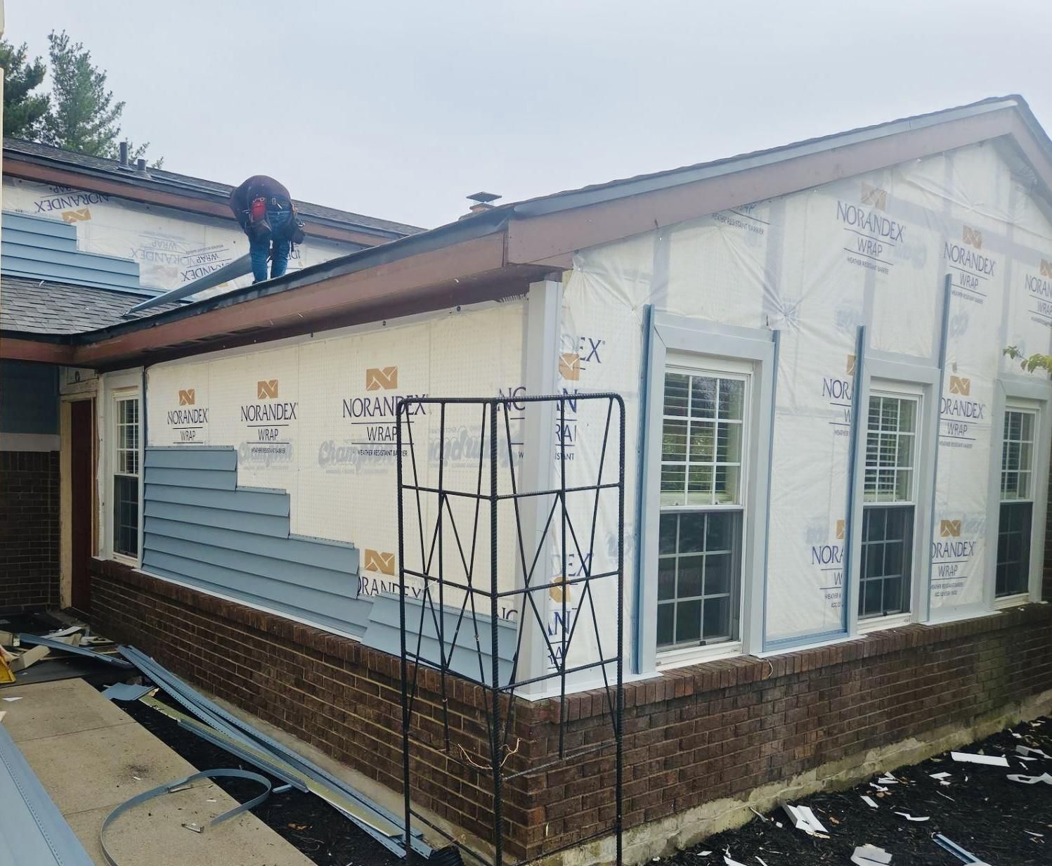 Blue vinyl siding being installed over house wrap on a brick ranch home, with a worker on the roof