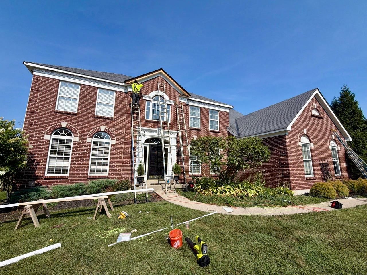 Fortress Solutions crew member on a ladder repairing fascia at the front gable of a red brick colonial, with new white trim boards on sawhorses