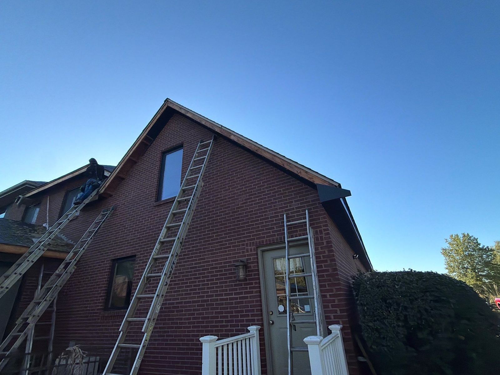 Crew member on a brick home roof edge replacing bare wood gable fascia and rake boards, with extension ladders against the house