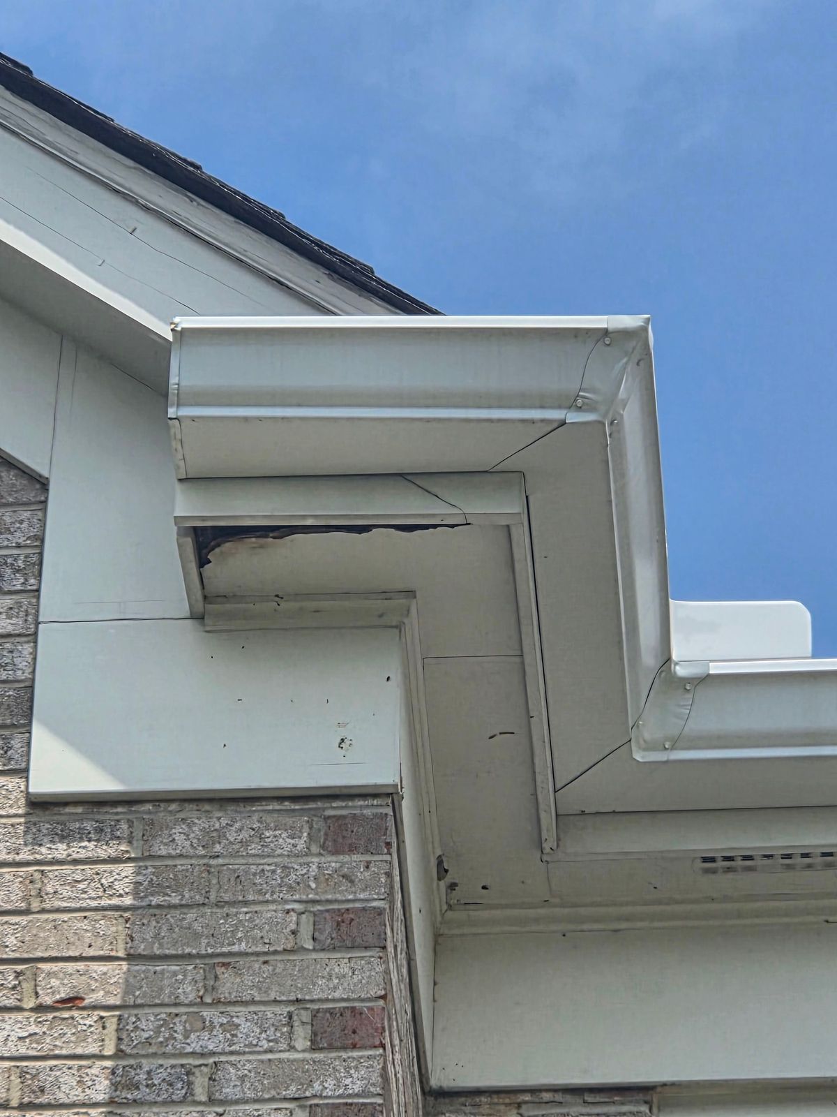 Before repair: damaged white soffit corner under a gutter on a brick home, with a missing section exposing wood underneath