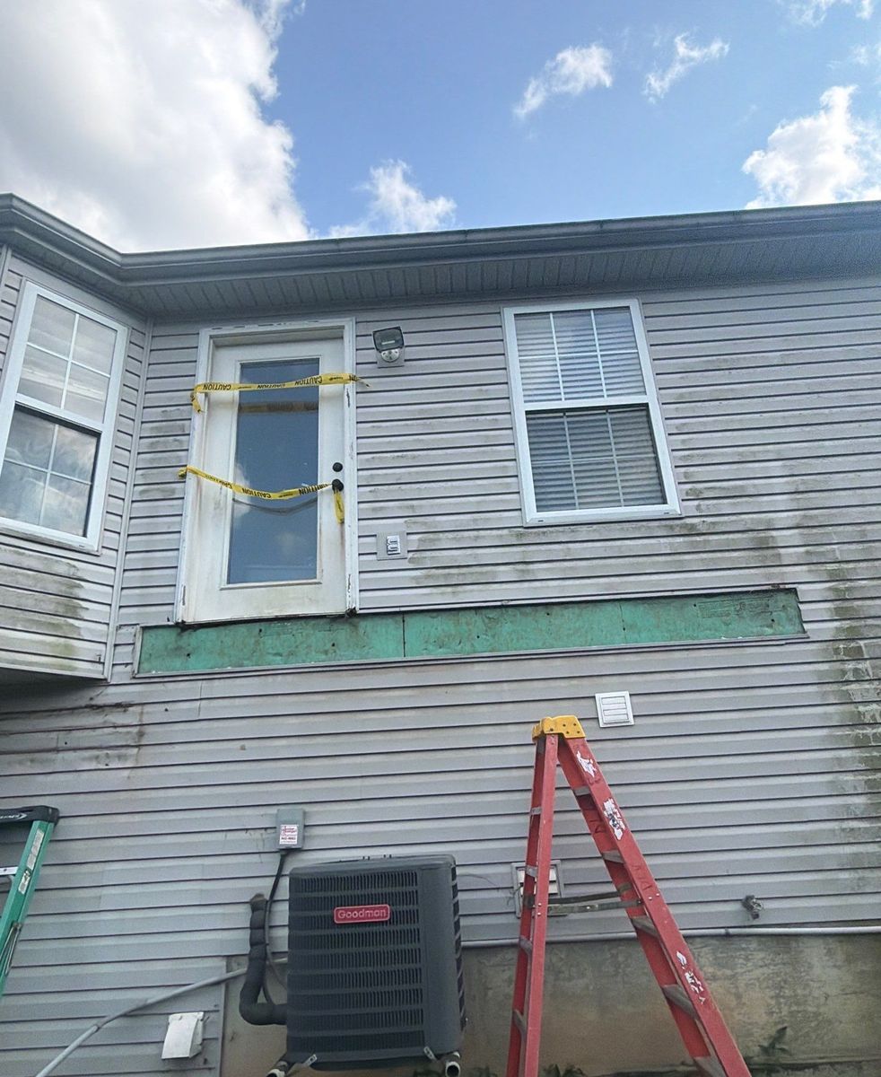 Back of a vinyl-sided home with siding removed and sheathing exposed where the deck ledger board will attach, door taped off for safety