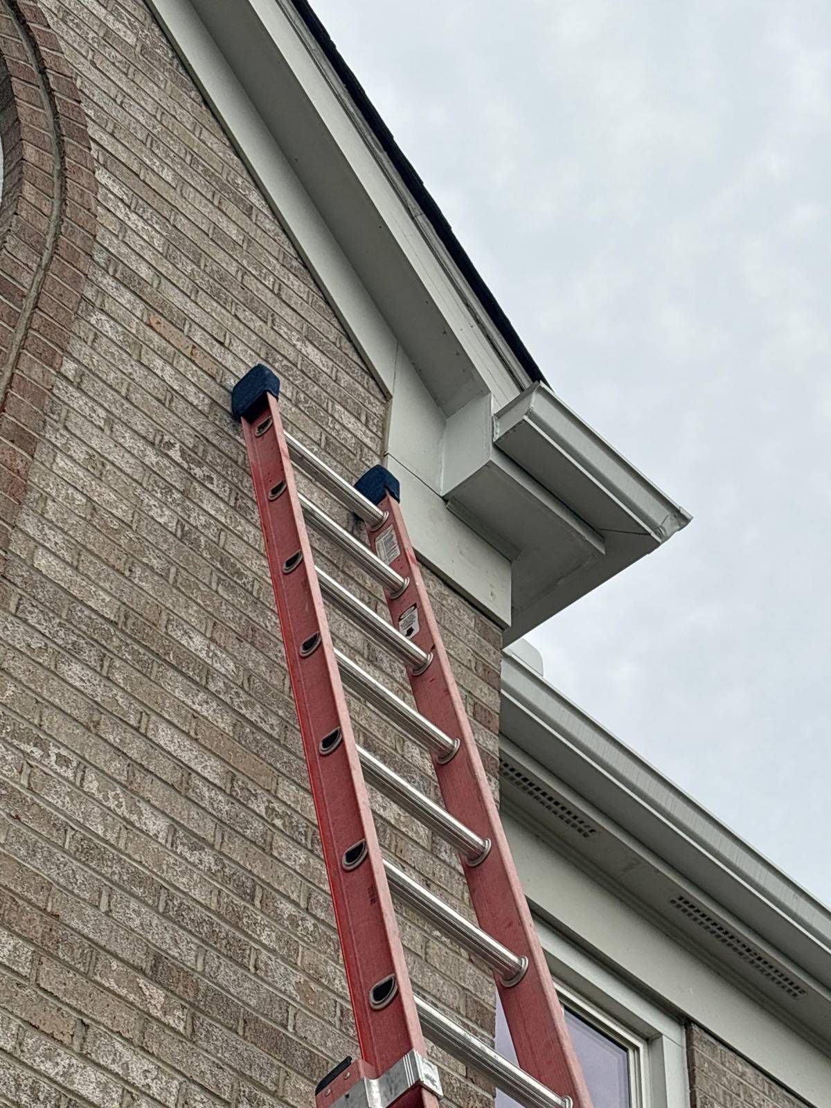 After repair: white soffit and fascia corner restored on a brick home, with a red extension ladder leaning against the wall
