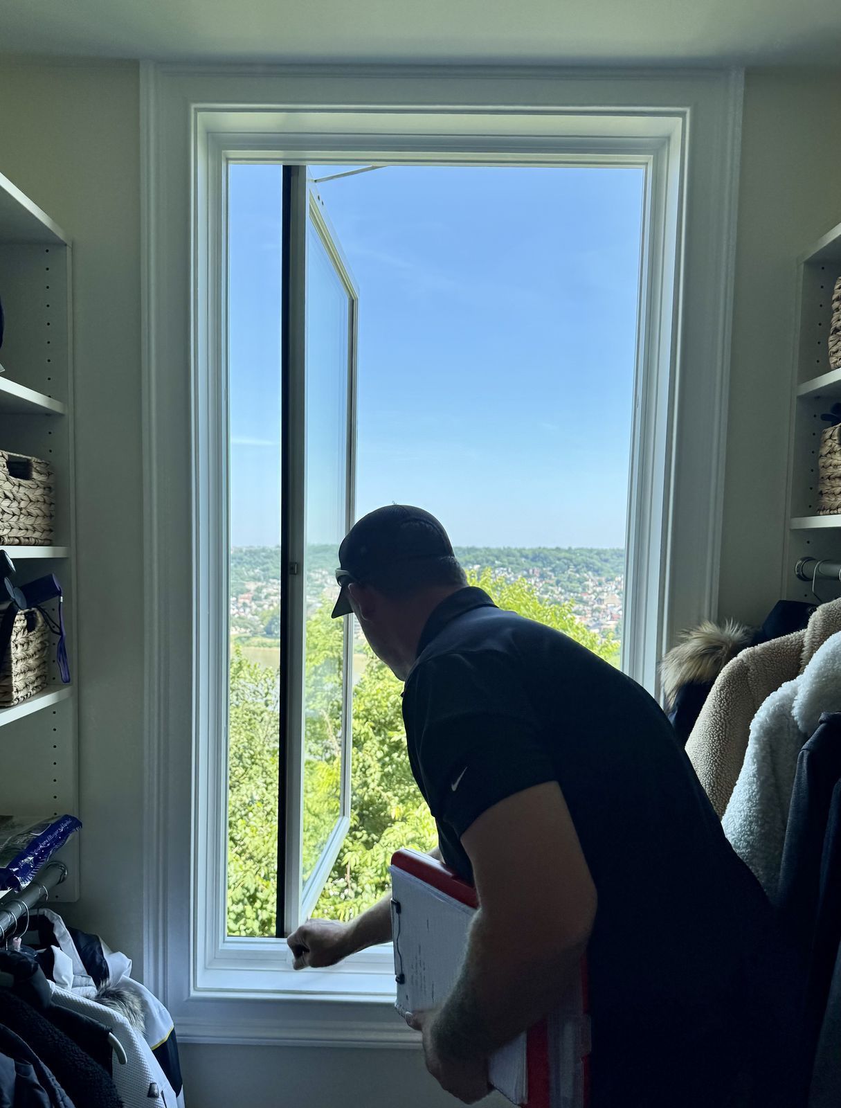 Man in a cap holding a clipboard opens a tall white casement window overlooking a wooded river valley