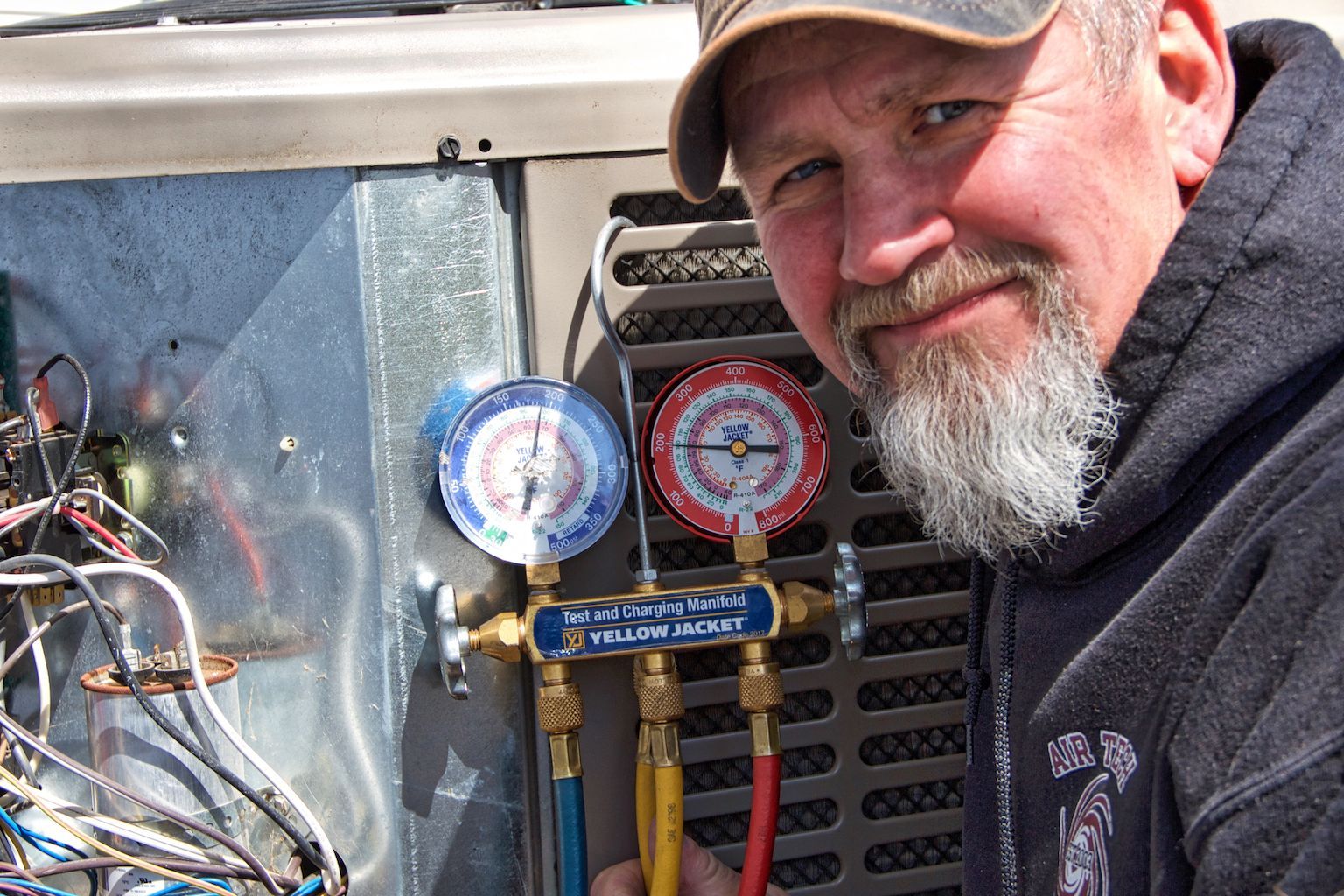 Man smiling beside HVAC gauges on a metal unit, with wires visible in the background.