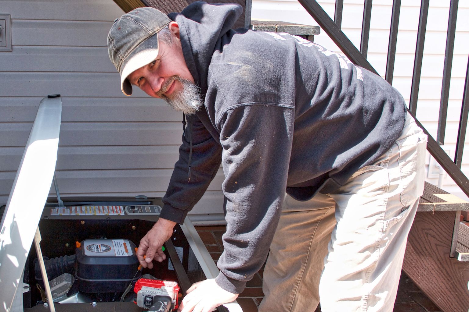 Man in a cap and gray hoodie working on equipment beside a house