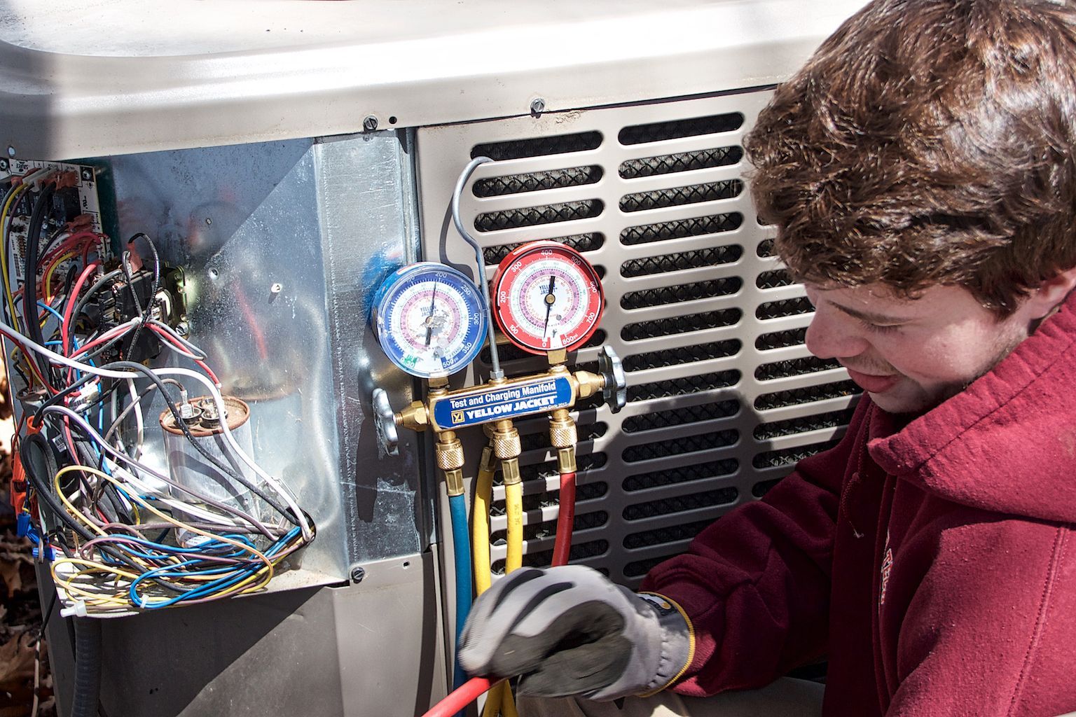 Technician using gauges to service HVAC unit with exposed wiring and vented panel