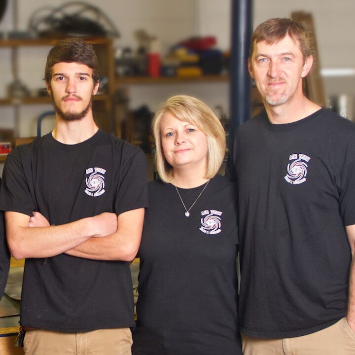 Three people in black shirts standing in a workshop, with shelves and equipment in the background.