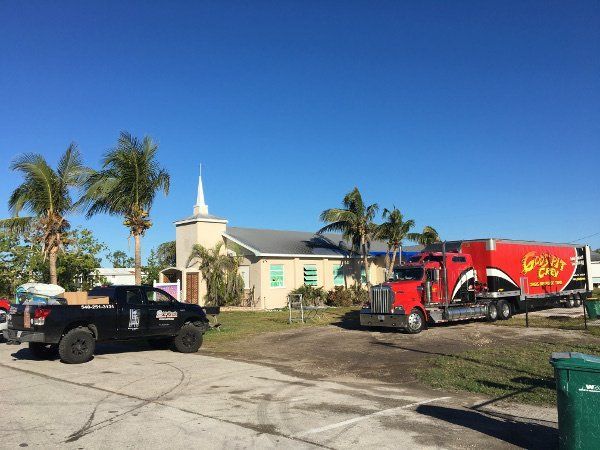 Two trucks parked outside a beige house with palm trees under a clear blue sky